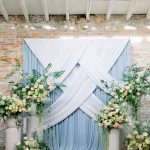 A wedding ceremony backdrop with intersecting white fabric draping over a light blue background, flanked by large floral arrangements with peach roses and greenery.