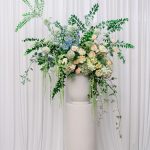 A beautiful ceremony floral pillar arrangement on a pedestal, with a mix of soft-colored flowers and greenery, standing before a draped backdrop