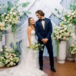 The happy couple celebrates their wedding day, standing before a beautiful backdrop of white and light blue draping and two large floral arrangements with peach roses and soft-toned flowers.