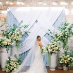 A stunning bride in her wedding dress, smiling as she stands in front of a white fabric and light blue draped backdrop with numerous floral arrangements on white pedestals.