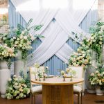 An elegant sweetheart table for two with a delicate floral arrangement and glassware, set in front of a white and light blue draped backdrop with large floral installations on either side.