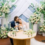 A newly married couple at their head table, with champagne glasses, in front of a beautifully draped backdrop and flowers at 128 South Event in Wilmington, NC.