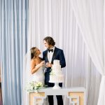 Newlyweds toast with champagne glasses next to their white wedding cake, with sheer draping in blue and white in the background at 128 South.