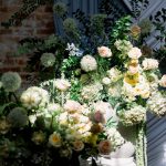 A close-up of wedding floral arrangements with blush and yellow roses, allium, and greenery, set against a backdrop of blue draping and exposed brick.