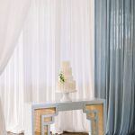 The wedding cake on display at 128 South, set in front of a backdrop of soft white and light blue draping, complementing the venue's elegant decor