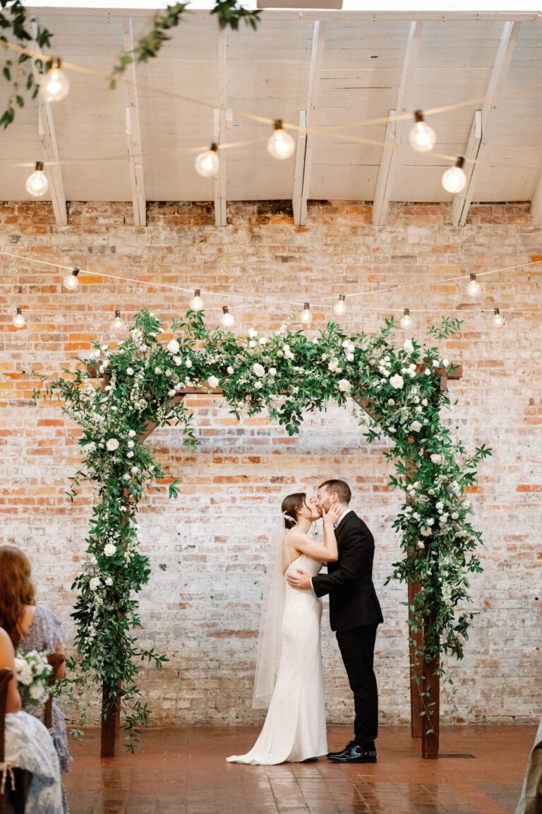 Couple under a greenery arch against the brick wall at Bakery 105